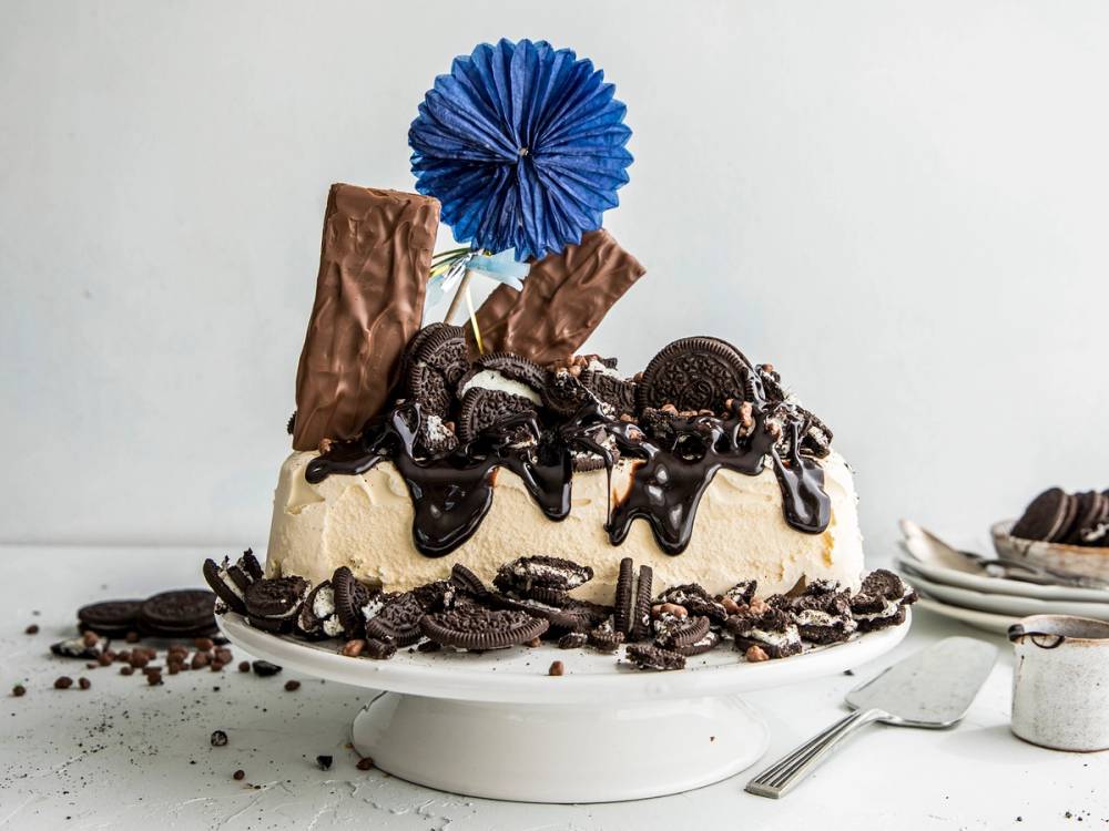 En Oreo-iskake pyntet med Oreo-kjeks, sjokoladesaus og en blå papirblomst på toppen, plassert på et hvitt fat.