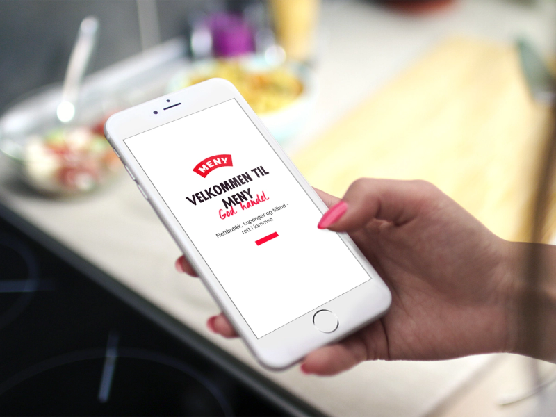 En person holder en smarttelefon som viser en velkomstskjerm for meny-handle-app med teksten 'Velkommen til', på et kjøkkenbord.