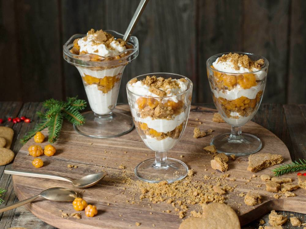 Tre glass med pepperkake-dessert med lag av krem, pepperkaker og karamellsaus på et trebord med julegrener og småkaker rundt.