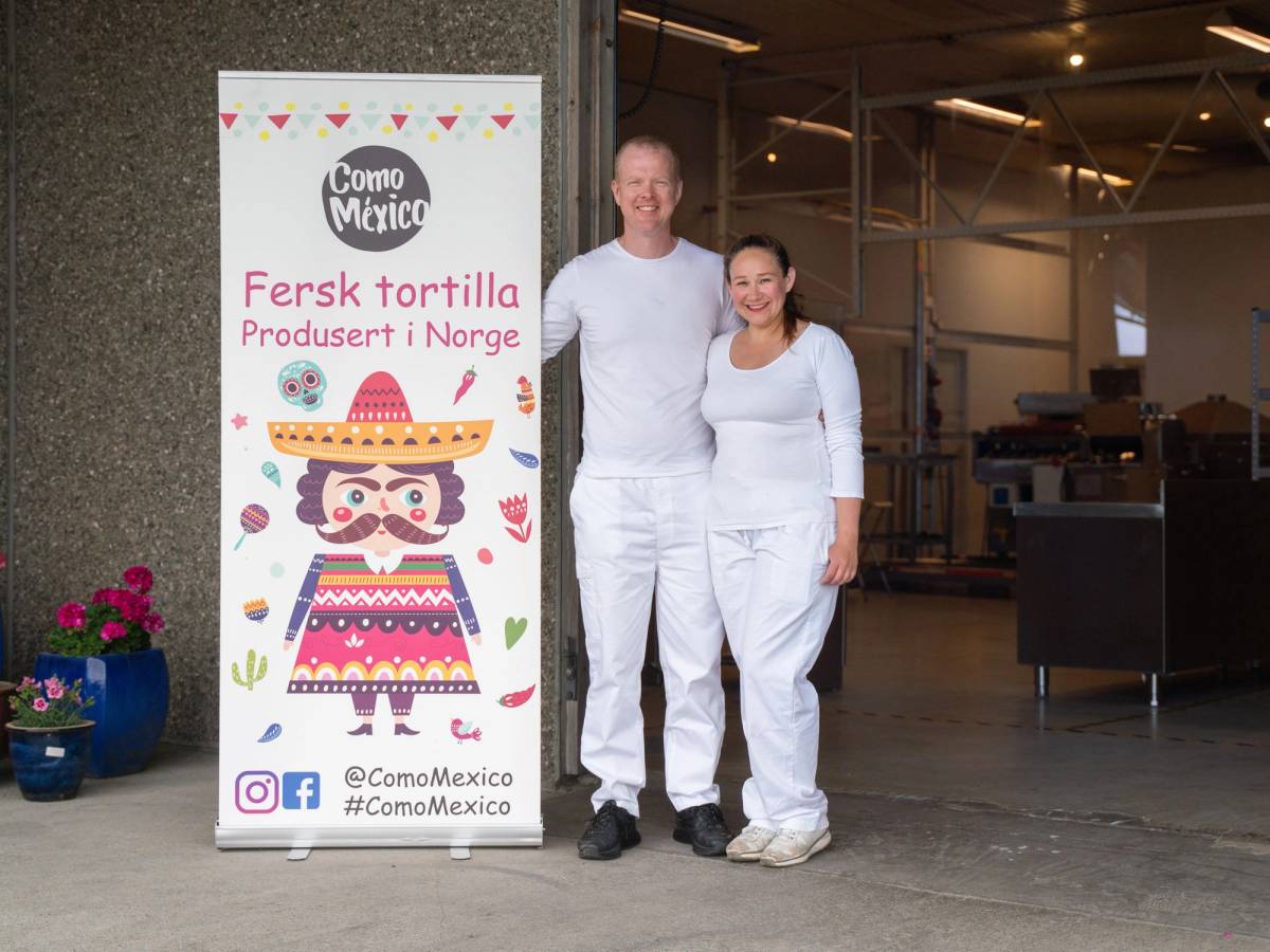 To personer står ved siden av en reklameskilt for fersk tortilla produsert i Norge, med en fabrikk i bakgrunnen.