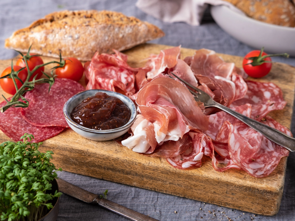 Frokostfat med spekemat, salamibiter, cherrytomater, og en liten skål med syltetøy på et tre- eller stekefat.