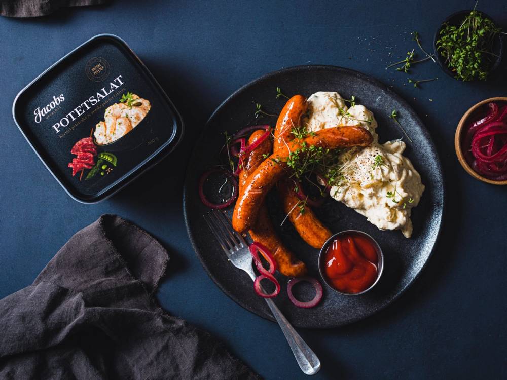 En tallerken med pølser, potetsalat og ketchup, med en boks Jacobs potetsalat i bakgrunnen.