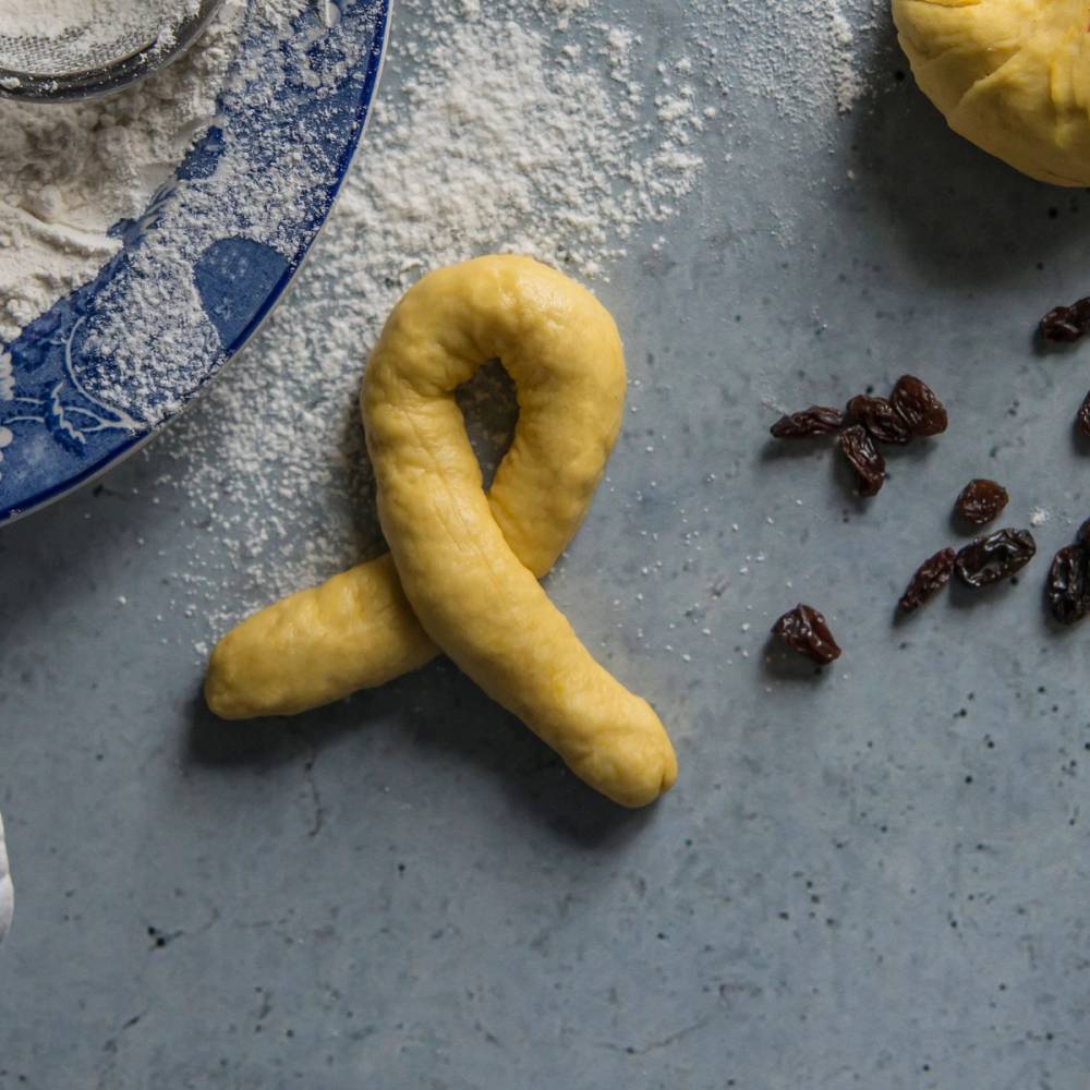 En lussekatt formet som en soldatgutt ligger på en melstrødd overflate med noen rosiner ved siden av.