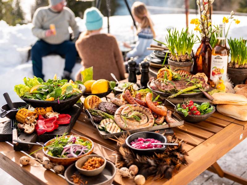 Grillmat med påskemat og grønnsaker på et utendørs bord med snø og mennesker i bakgrunnen.