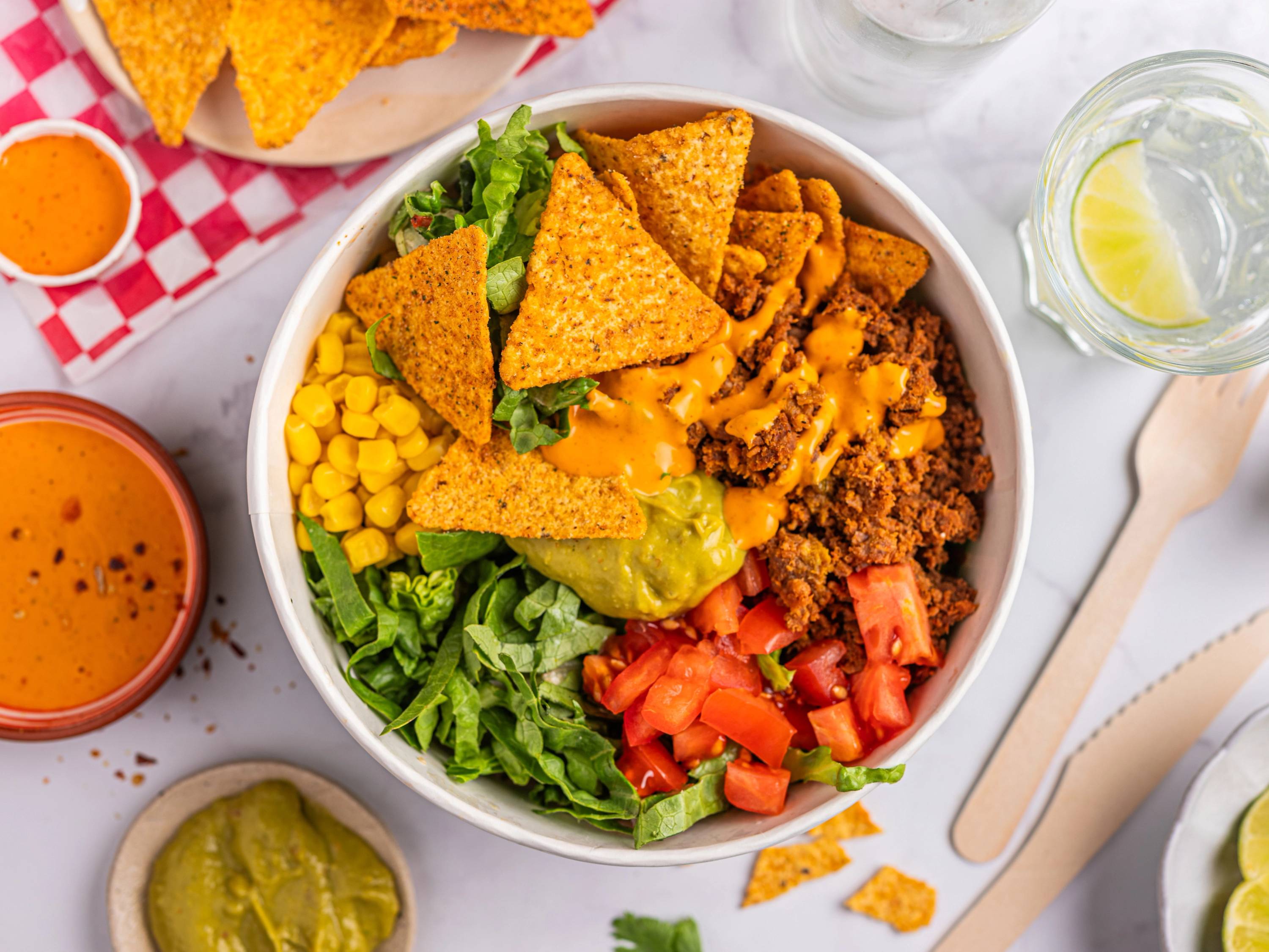 En bolle med salat og ulike tilbehør som tortillachips, mais, tomater og guacamole, omgitt av små skåler med saus og en glass med sitronvann.