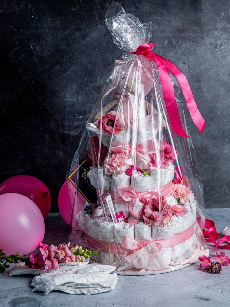 En bleiekake dekorert med rosa og hvite elementer, pakket inn i gjennomsiktig cellofan med rosa sløyfe, omgitt av rosa ballonger og blomster.
