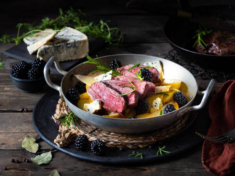 Stekt kjøtt med bær og urter på en tallerken, omgitt av friske bær og krydderurter på et mørkt bord.