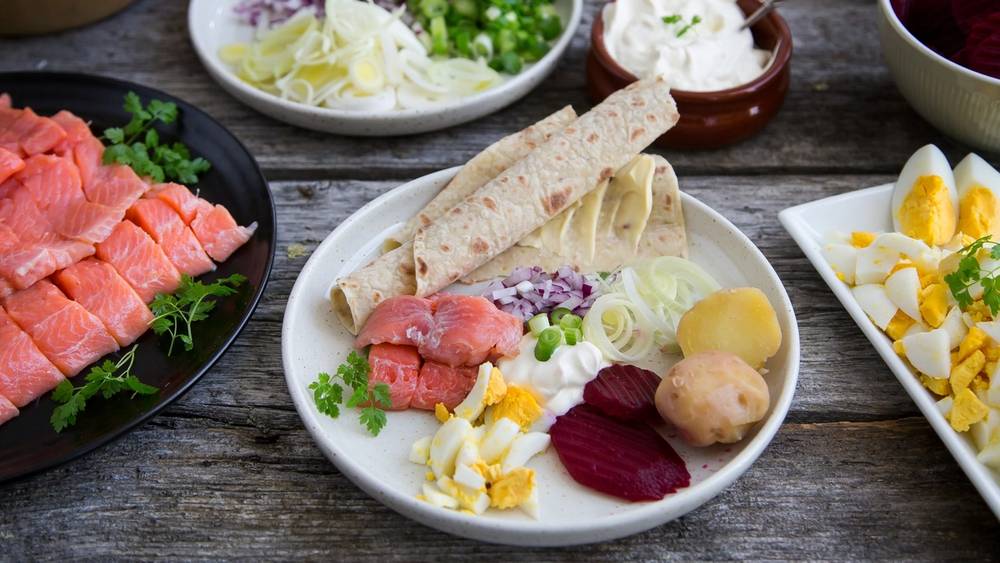 Tradisjonell rakfisk med tilbehør som flatbrød, løk, rømme, poteter og sylteagurk på et bord.