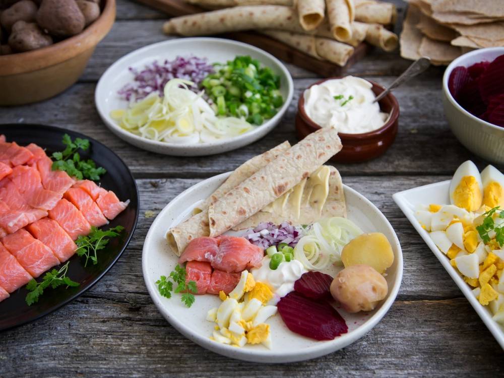 Tradisjonell rakfisk med tilbehør som flatbrød, løk, rømme, poteter og sylteagurk på et bord.