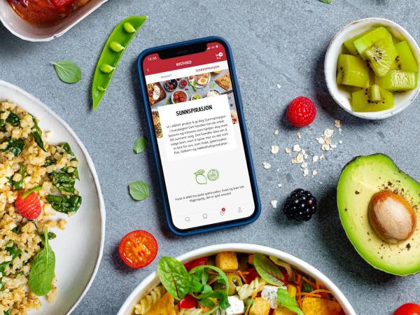 En mobiltelefon som viser en nettside om sunnspirasjon, omgitt av frukt og grønnsaker på en bordflate.