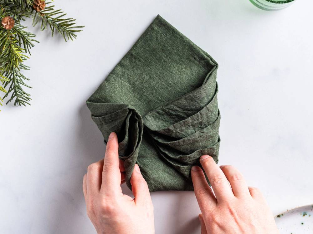 En person folder en grønn serviett til å ligne et juletre, med noen grener av gran på venstre side.