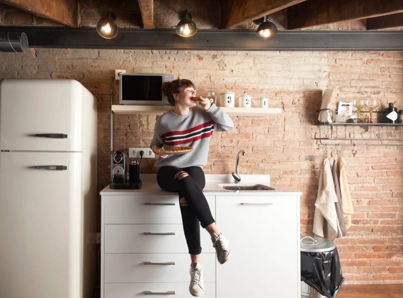 En student som drikker fra et glass på kjøkkenet, med hvite skuffer og en hvit kjøleskap i bakgrunnen.