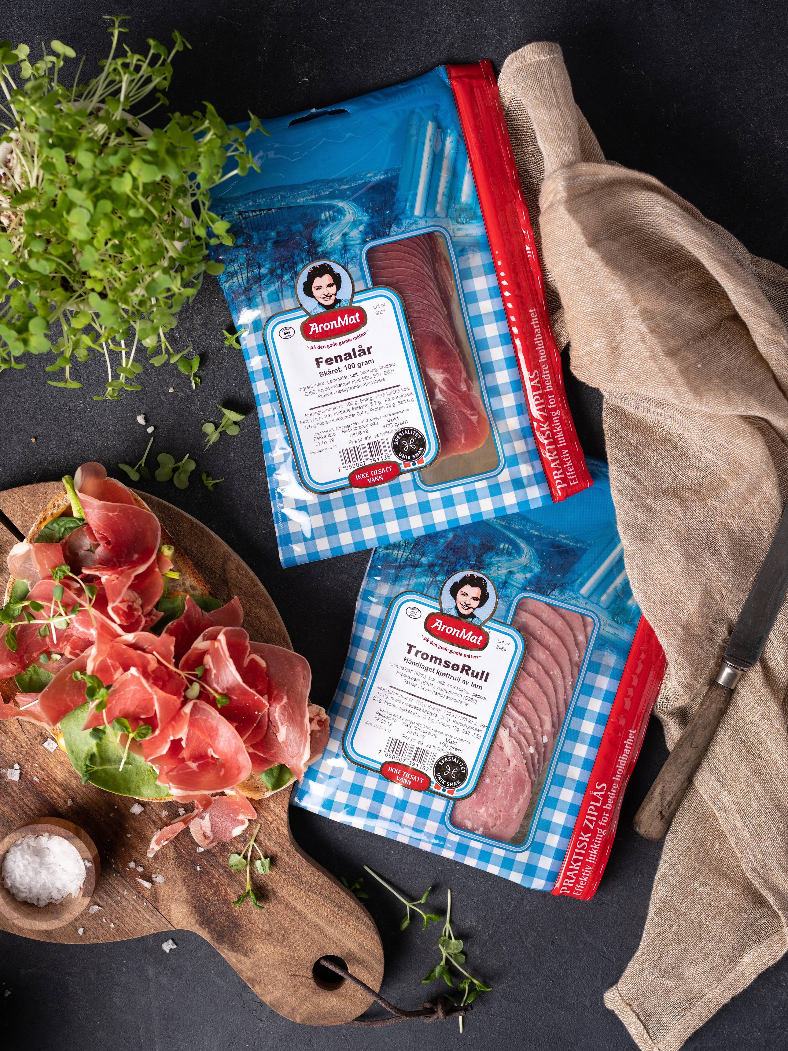 To pakker med ferdigskåret kjøttpålegg fra Aron, plassert på et kjøkkenbord med en skinke og urter i nærheten.