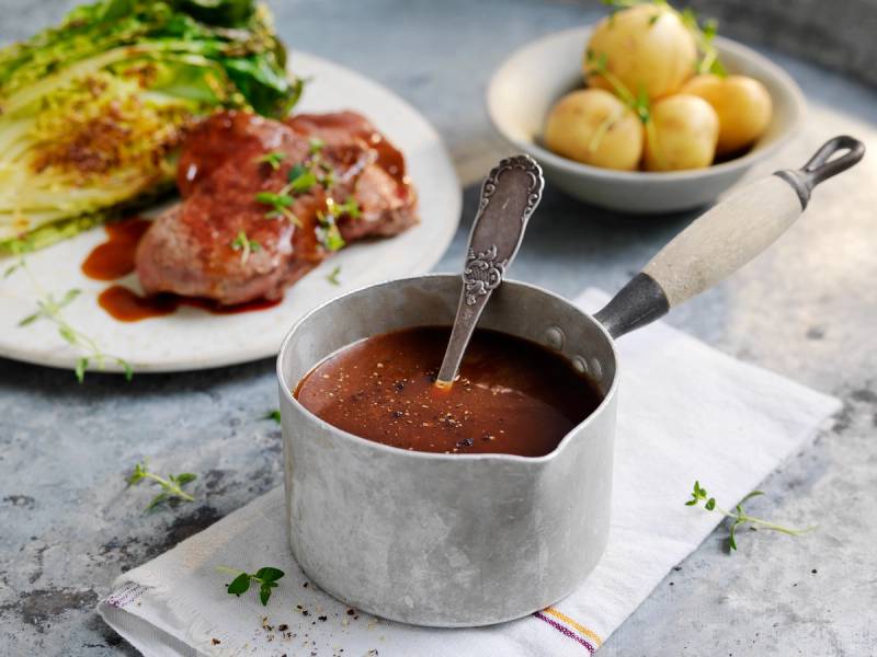 Rødvinssaus i en liten kjele med en skje oppi, servert med en tallerken med kjøtt og grønnsaker i bakgrunnen