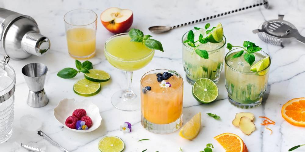 Et utvalg av fargerike mocktails med sitrusfrukter og mynte på et hvitt bord, omgitt av barutstyr og frukt.