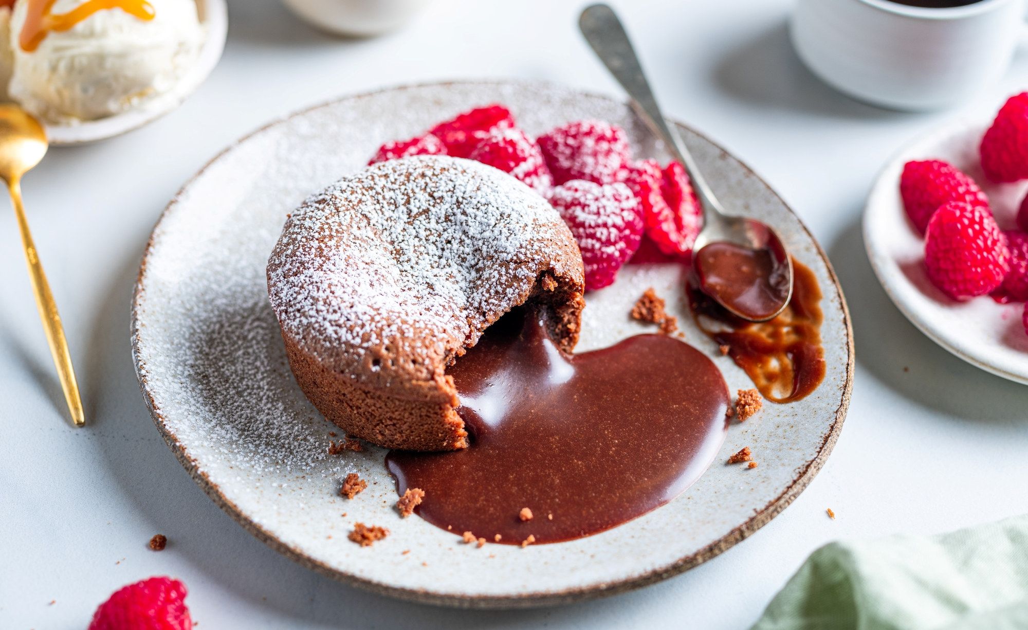 Sjokoladefondant med flytende sjokoladekjerne på en tallerken, omgitt av friske bringebær og melis, med en kopp kaffe i bakgrunnen.