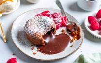 Sjokoladefondant med flytende sjokoladekjerne på en tallerken, omgitt av friske bringebær og melis, med en kopp kaffe i bakgrunnen.