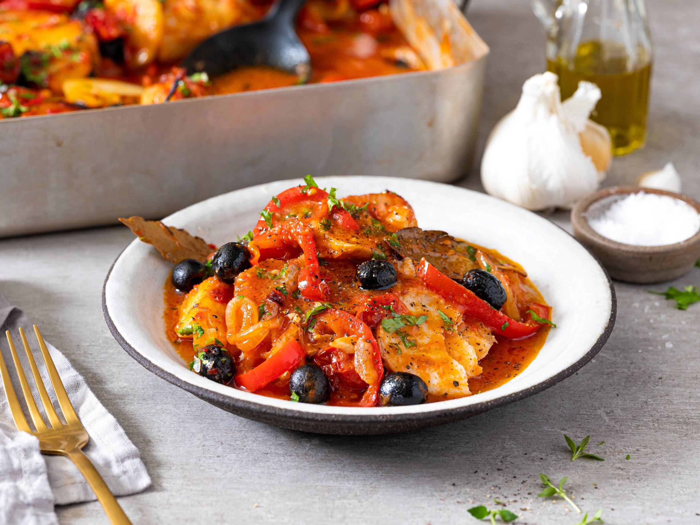 Bacalao med tomater, paprika og oliven på en hvit tallerken, med hvitløk og olivenolje i bakgrunnen.