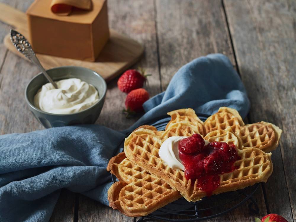 Vafler med jordbær og krem på en tallerken, med en skål med yoghurt og noen jordbær i bakgrunnen.
