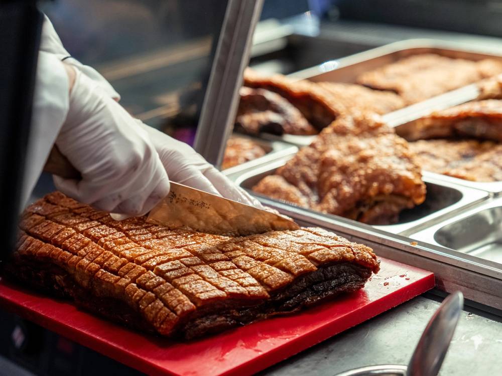 En person skjærer opp store stykker av grillet kjøtt i en matbutikk eller på en kjøttbutikk, med flere kjøttstykker i bakgrunnen.