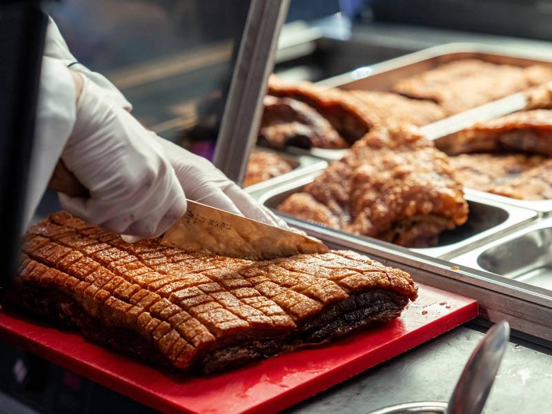 En person skjærer opp store stykker av grillet kjøtt i en matbutikk eller på en kjøttbutikk, med flere kjøttstykker i bakgrunnen.