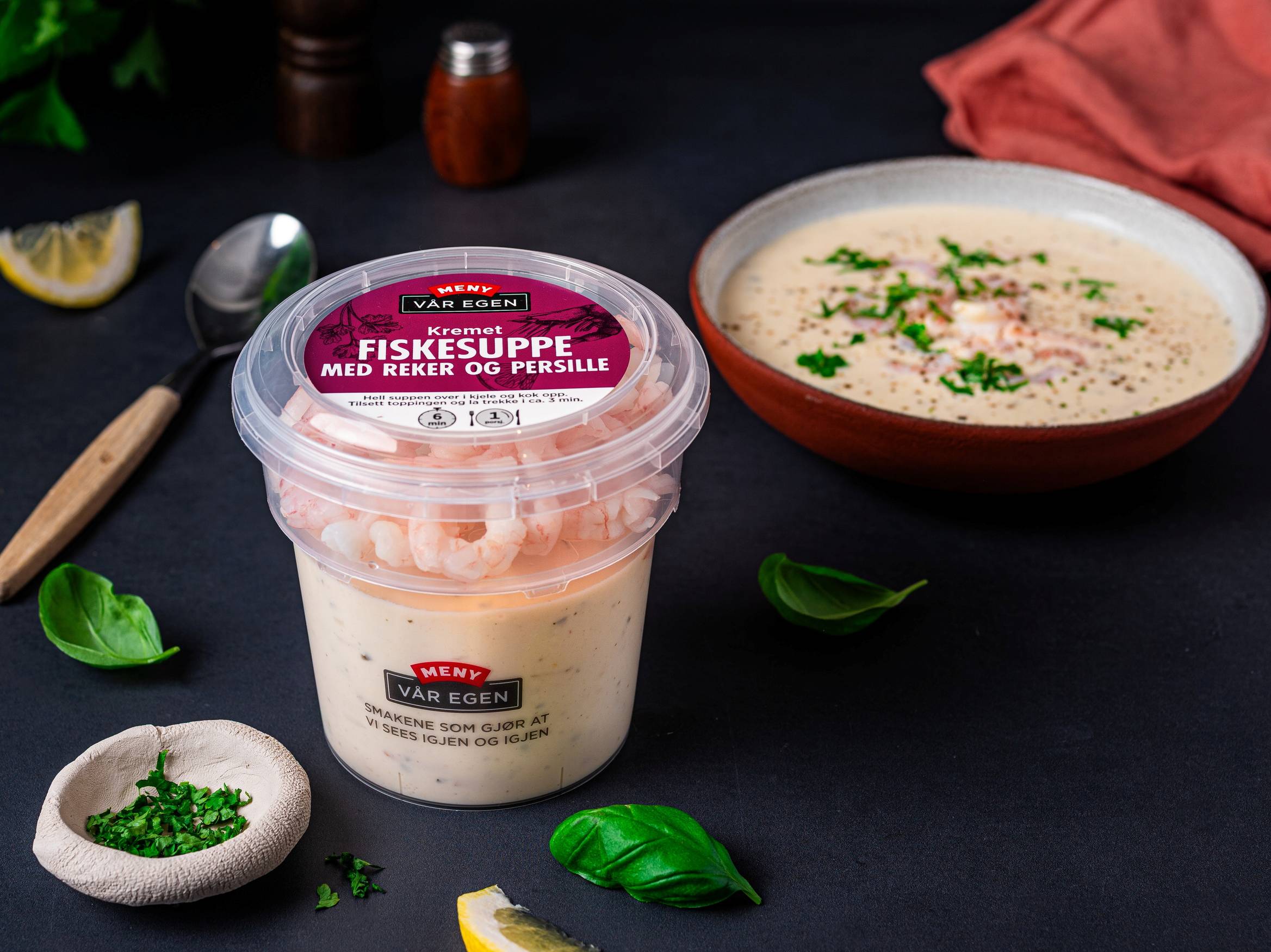 Fiskesuppe med reker og persille i en hvit bolle, omgitt av sitronbiter, urter og krydder på en svart bakgrunn.