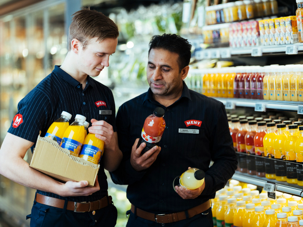 To ansatte i butikk som sammen vurderer og velger varer på hyllen, med en kurv med drikkevarer i hendene.