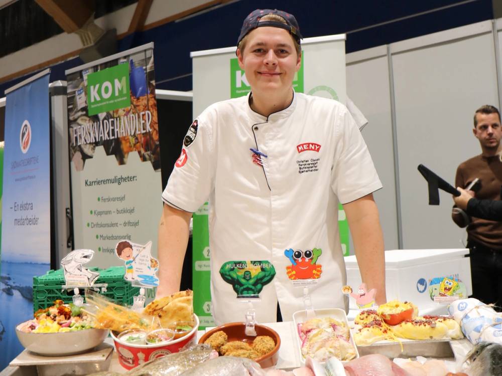 En person står bak et bord med ulike sjømatretter og kaker, og holder en gullmedalje, på en utstilling eller konkurranse.