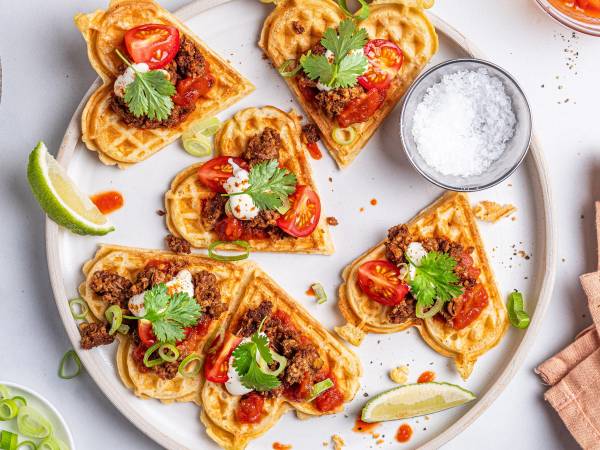 Vaffel nachos med tomatbiter, koriander og limebåter på en hvit tallerken, med en liten skål salt og en liten skål med saus i nærheten.