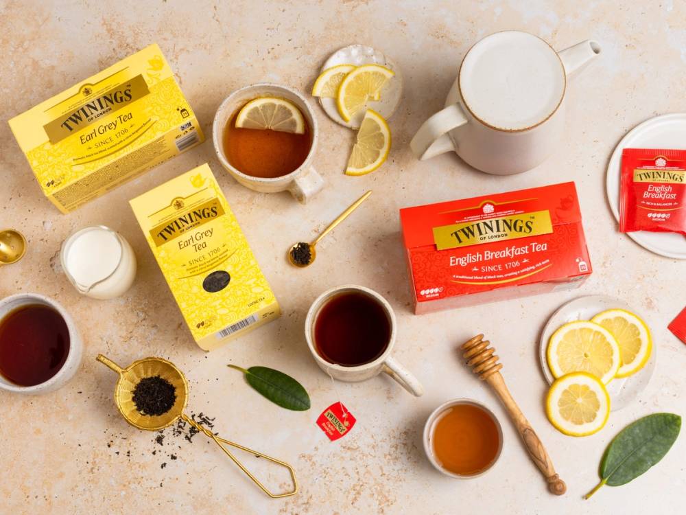 Ulike typer te, sitronbåter, en kopp med te, og honning på et bord, med Twinings tepakker i bakgrunnen.