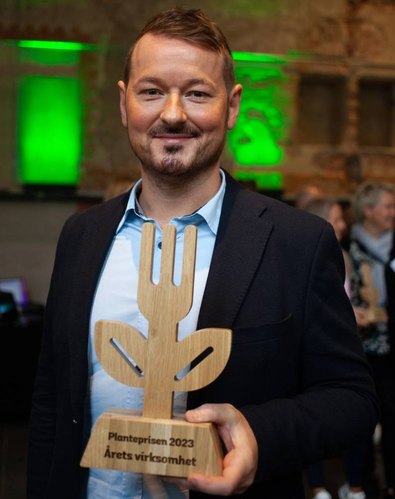 En mann i dress holder en treutskåret planteprisen 2023 med teksten 'Årets virksomhet' på en plakat foran seg.