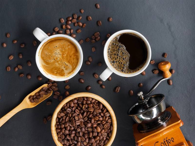 To kopper med kaffe, en skål med kaffebønner, og en kaffekvern på en mørk bakgrunn.