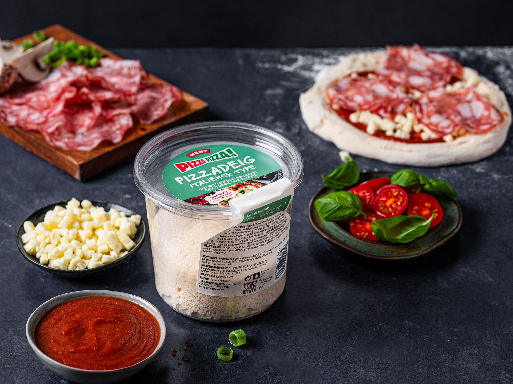 Ulike ingredienser for å lage pizza, inkludert skinke, mozzarellaost, cherrytomater og pizzasaus, er arrangert på en svart overflate.