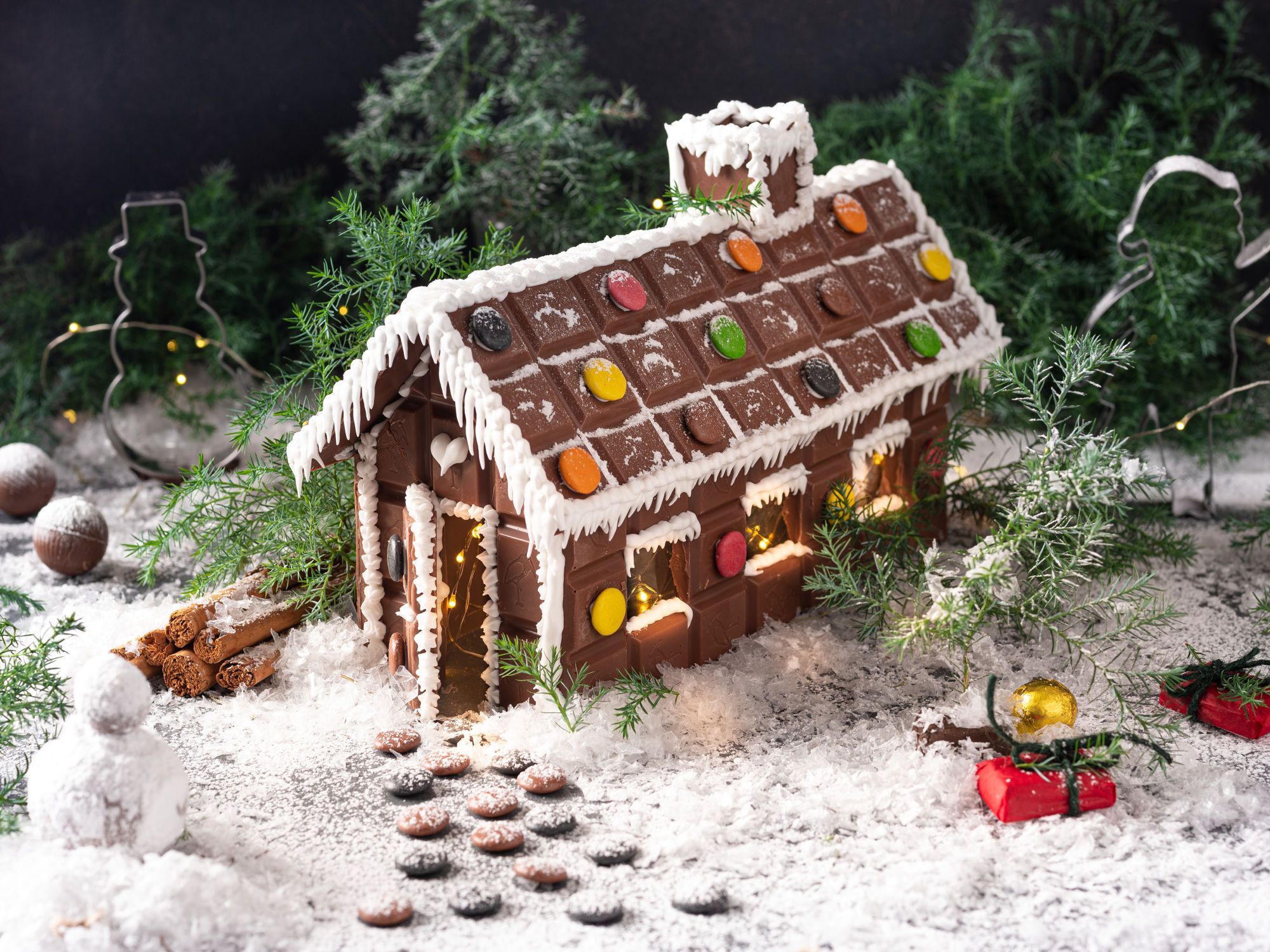 Et juleinspirert sjokoladehus dekorert med fargerike godteri og hvit glasur, plassert på snøaktig underlag med julepynt rundt.