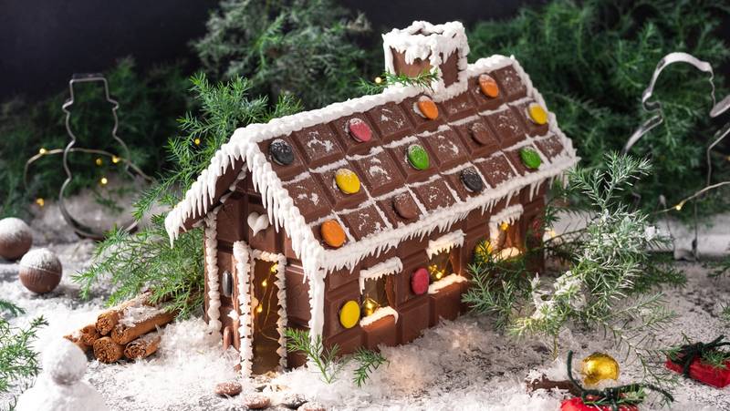 Et juleinspirert sjokoladehus dekorert med fargerike godteri og hvit glasur, plassert på snøaktig underlag med julepynt rundt.