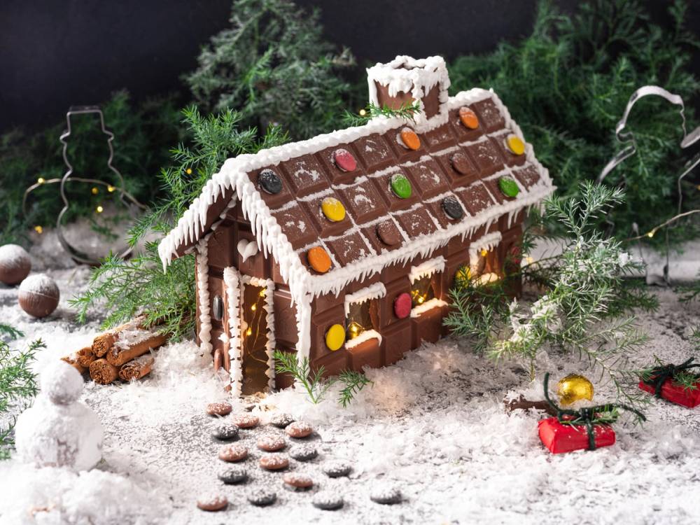 Et juleinspirert sjokoladehus dekorert med fargerike godteri og hvit glasur, plassert på snøaktig underlag med julepynt rundt.