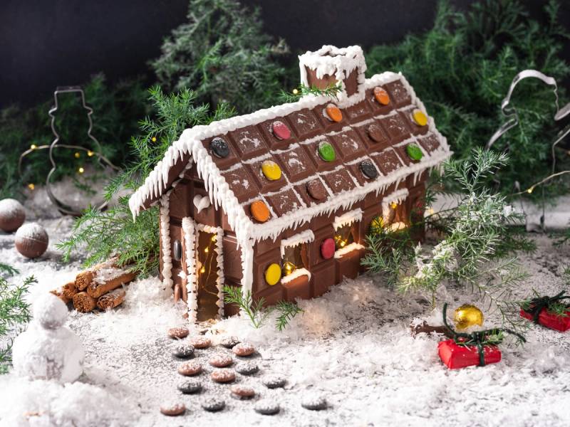 Et juleinspirert sjokoladehus dekorert med fargerike godteri og hvit glasur, plassert på snøaktig underlag med julepynt rundt.