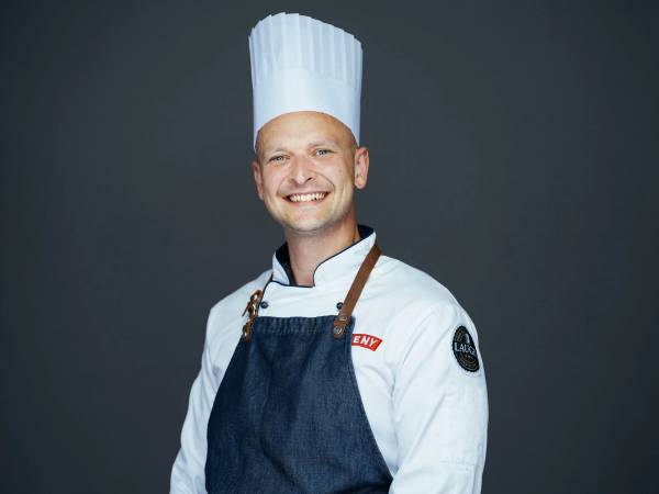 En person kledd som kokk med hvit kokkehatt og hvit jakke, iført en blå forkle, mot en mørk bakgrunn.