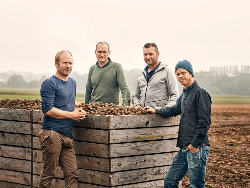 Fire personer står rundt en stor trebeholder fylt med poteter på en jordete åker, med åker og bygninger i bakgrunnen.
