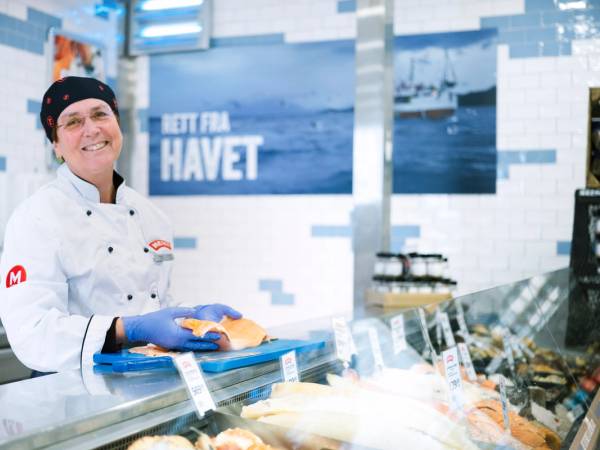 En person i hvit kokkeuniform står bak en fiskedisk med fersk sjømat i en butikk eller fiskebutikk, med en blå og hvit bakgrunn.