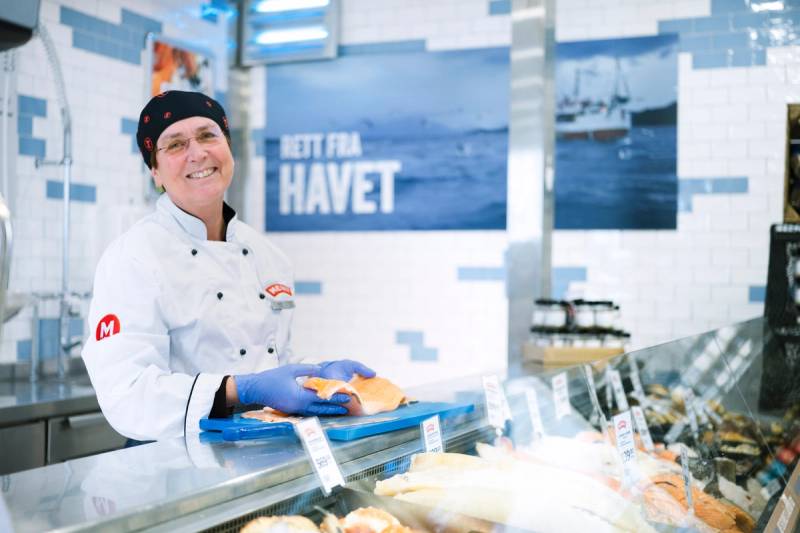 En person i hvit kokkeuniform står bak en fiskedisk med fersk sjømat i en butikk eller fiskebutikk, med en blå og hvit bakgrunn.