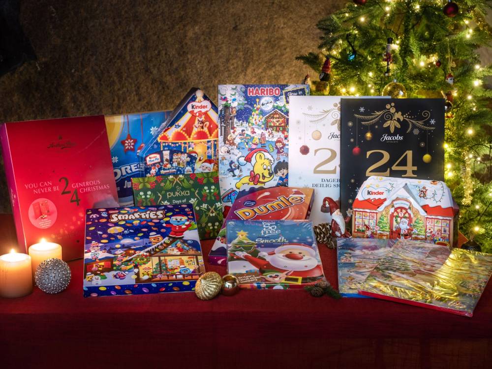 Ulike julekalendere og gaver er plassert foran et juletre med lys og pynt, på en borddekket overflate.
