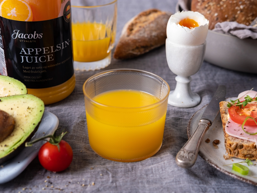 Frokostbord med glass med appelsinjuice, brød, et kokt egg, avokado og en tomat.