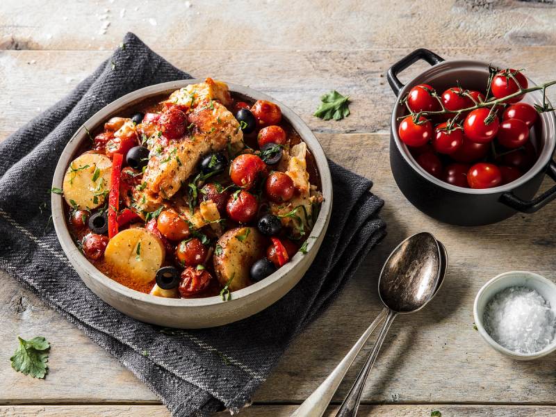 Bacalao med poteter, cherrytomater og urter i en gryte, med en skje og en liten bolle med salt ved siden av, på et trebord.