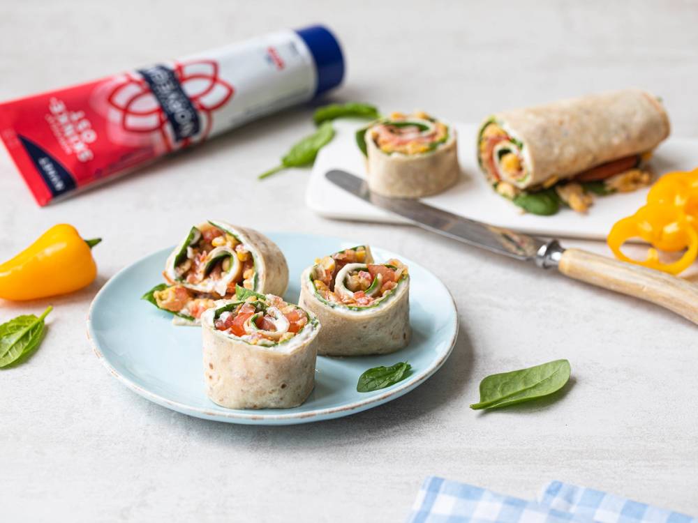 Lefseruller med smøreost og grønnsaker på en hvit tallerken, med en tube smøreost og en kniv i bakgrunnen.