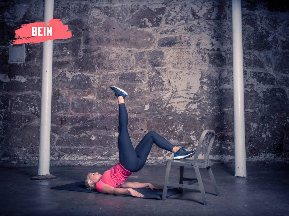 En person utfører et ettbeins rompeløft med en stol som støtte, på en treningsmatte foran en murvegg.