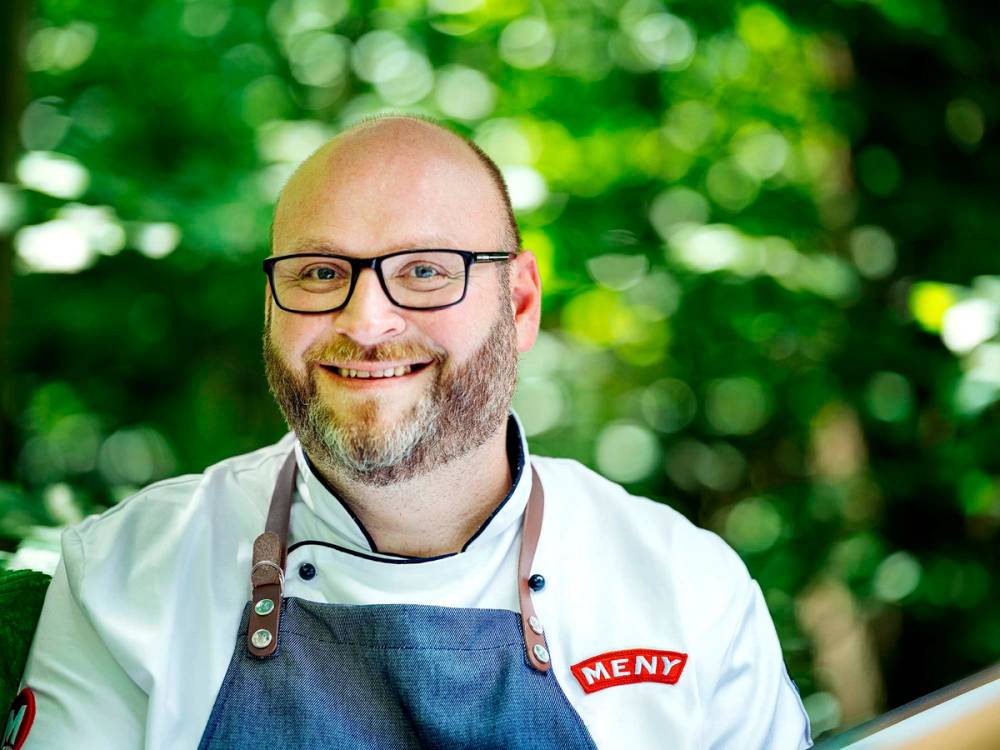 En person iført kokkeuniform og forkle står utendørs med grønt løv i bakgrunnen, og har ansiktet dekket med en blur-effekt.