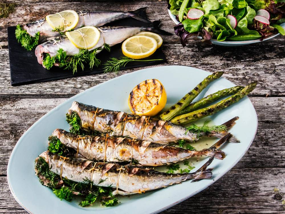 Grillet makrell med sitronbåter og grillede asparges på et hvitt fat, med en salat og sitroner i bakgrunnen.