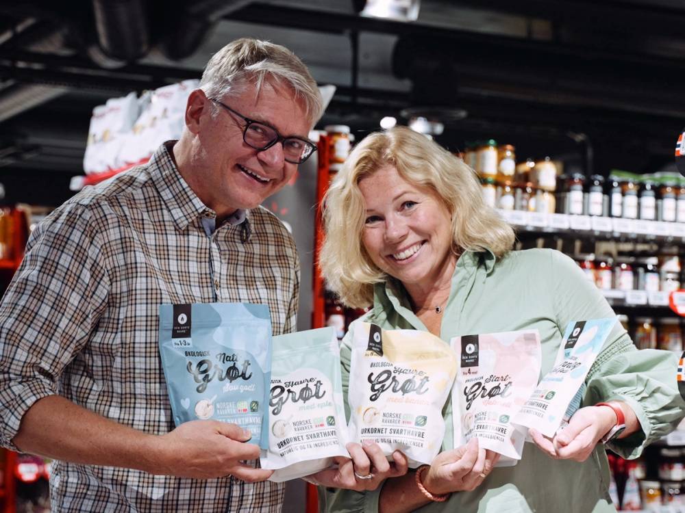 To personer holder opp poser med havreprodukter i en butikk, med hyller fylt av ulike varer i bakgrunnen.