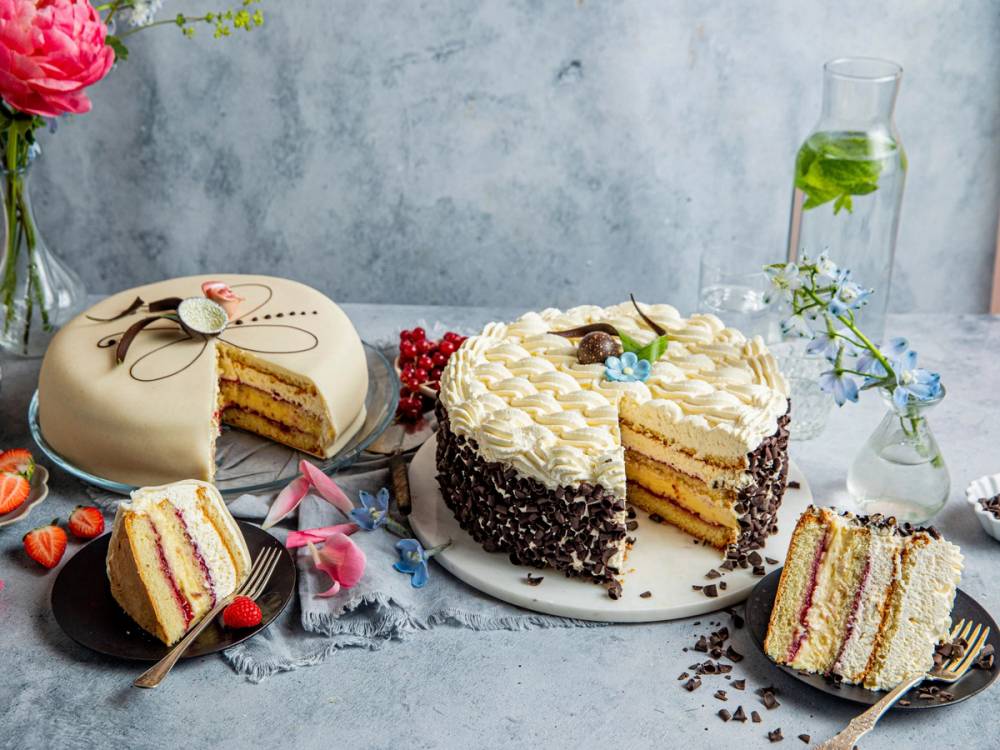 En bløtkake med marsipanlokk og pyntet med blomster og bær, med flere kakeskiver på siden.